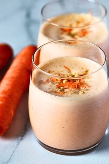 Two glasses of carrot cake smoothie with grated carrot topping and fresh carrots on a marble surface