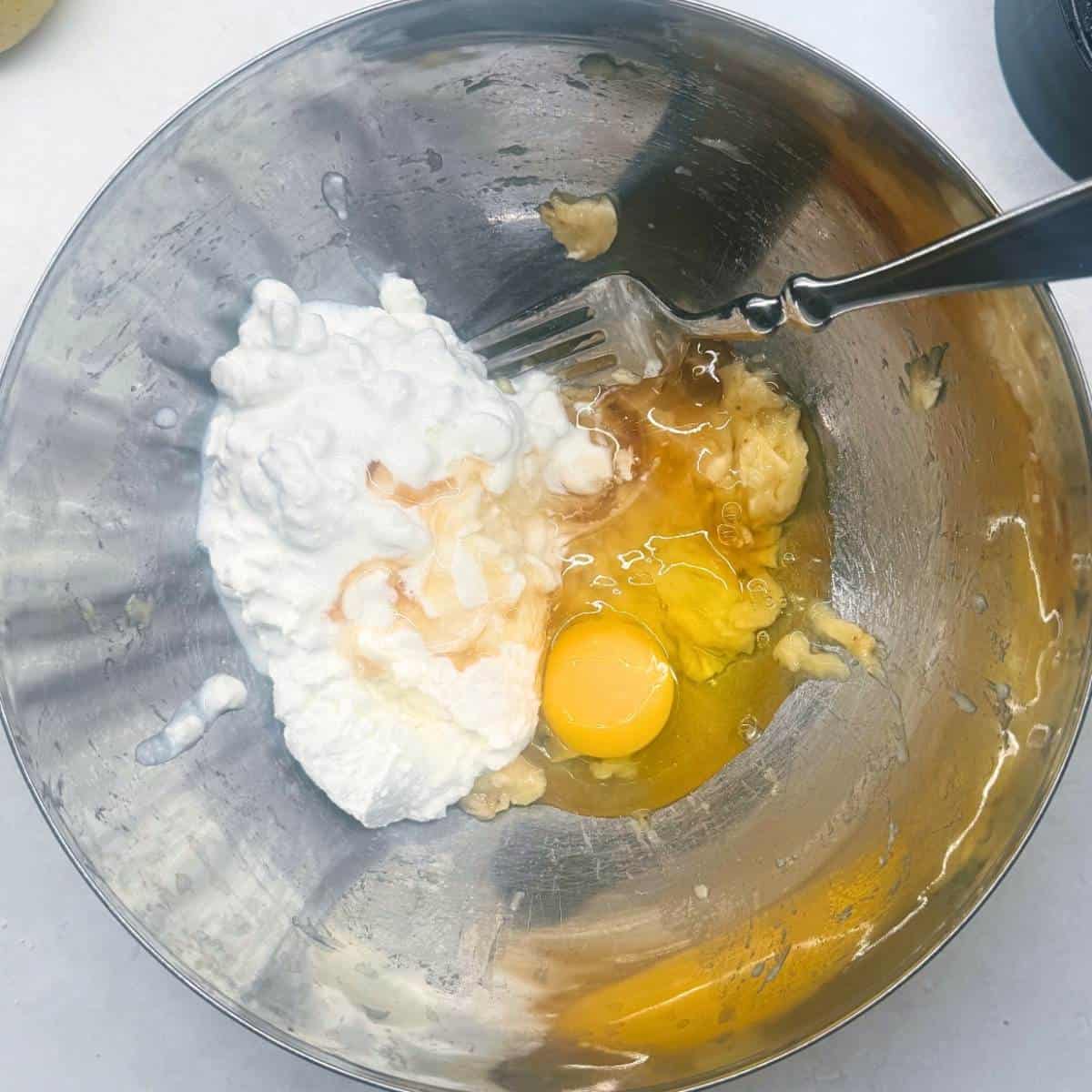 Mixing cottage cheese, egg, and mashed banana in a metal bowl with a fork